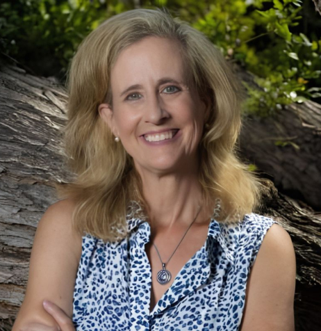 Woman named Melissa Brymer with long blonde hair wearing a blue-dotted white sleeveless blouse smiling in front of a tree.