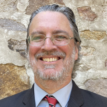 Portrait of Kenneth Kinter with gray hair, beard, and glasses in a suit and tie with a rock wall in the background