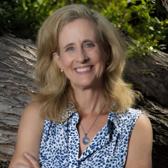 Woman named Melissa Brymer with long blonde hair wearing a blue-dotted white sleeveless blouse smiling in front of a tree.